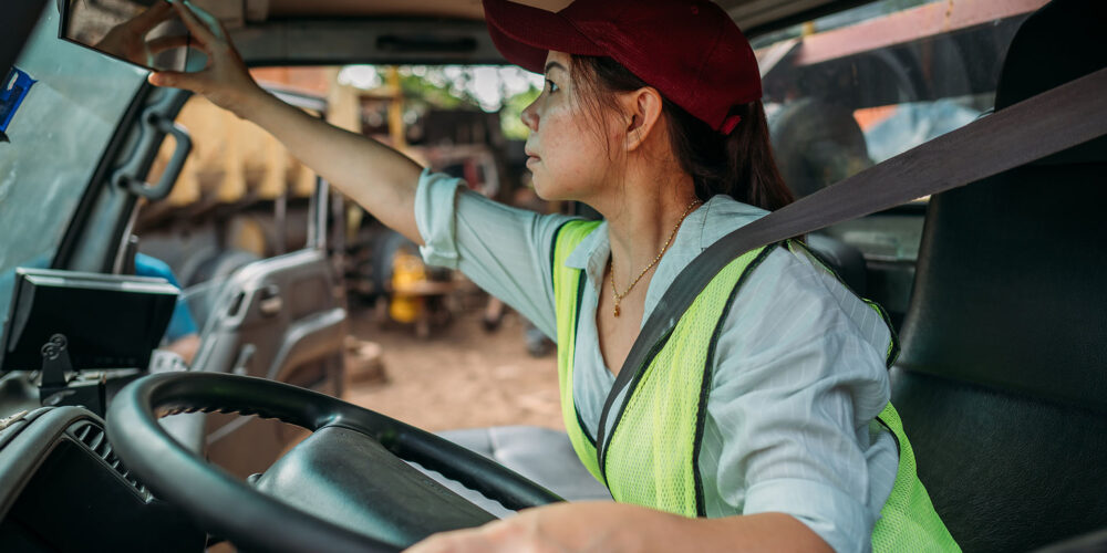 Personal perspective of mid 30s woman in casual clothing, cap, and reflective vest sitting in driver’s seat, adjust mirror and looking straight ahead, ready to begin road trip.