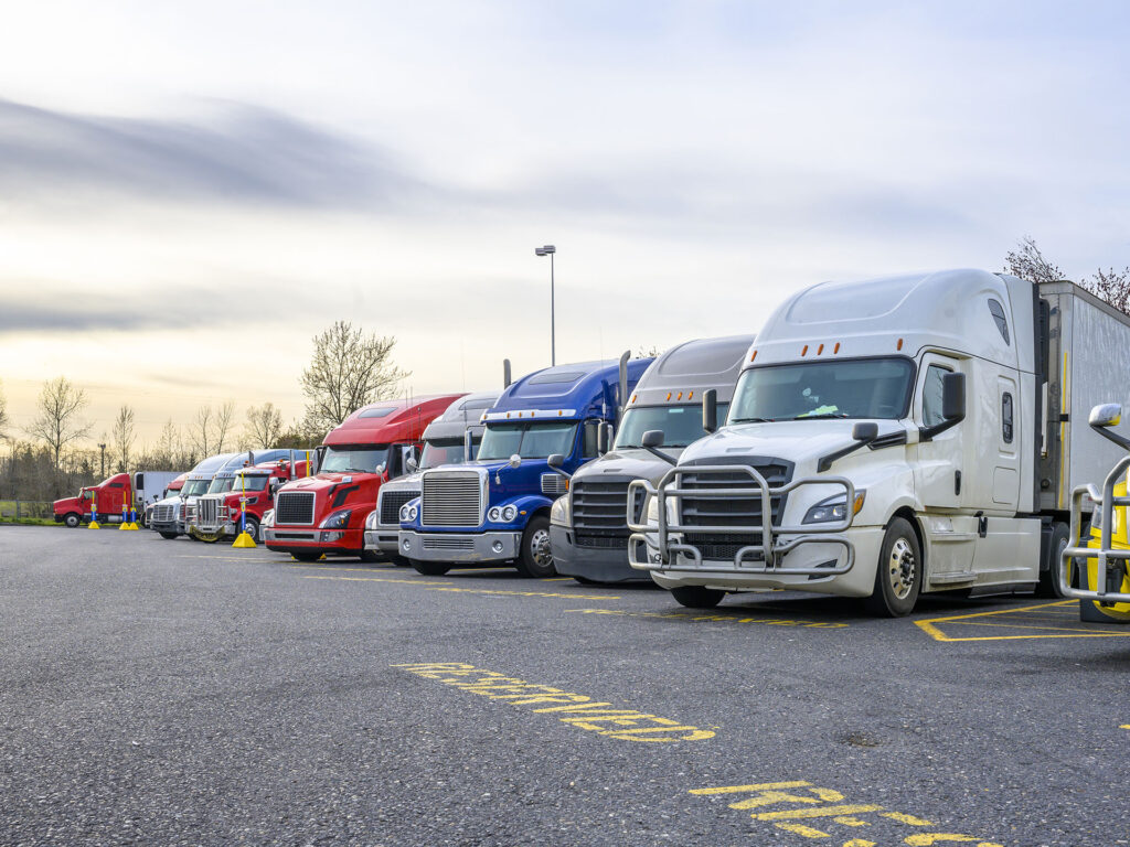 Different big rigs semi trucks with semi trailers standing in row on truck stop parking lot with reserved spots for truck driver rest and compliance with established truck driving regulations