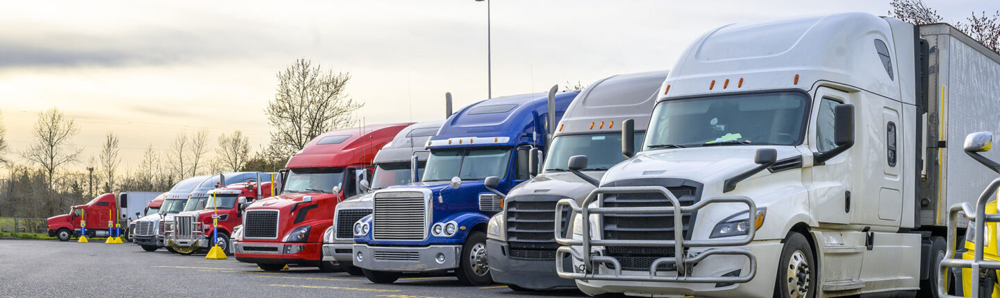 Different big rigs semi trucks with semi trailers standing in row on truck stop parking lot with reserved spots for truck driver rest and compliance with established truck driving regulations