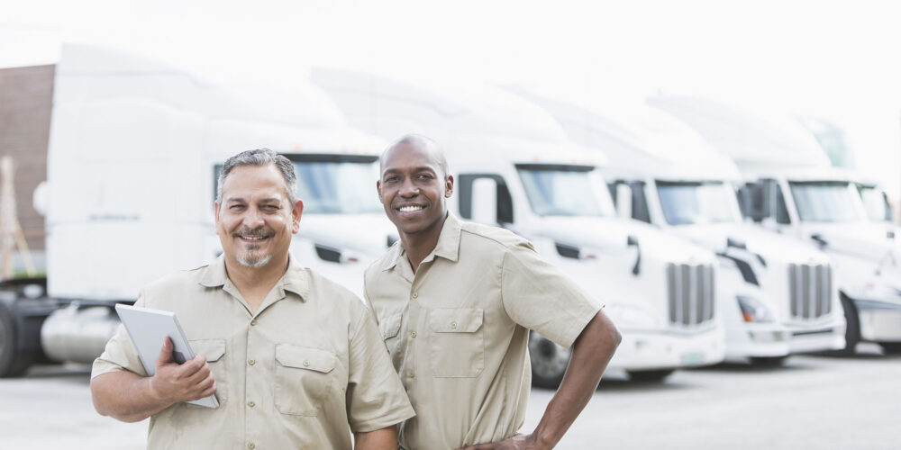 Two multi-ethnic mature men working at a trucking company. They are smiling at the camera, standing in front of a row of parked semi-trucks at a distribution warehouse.