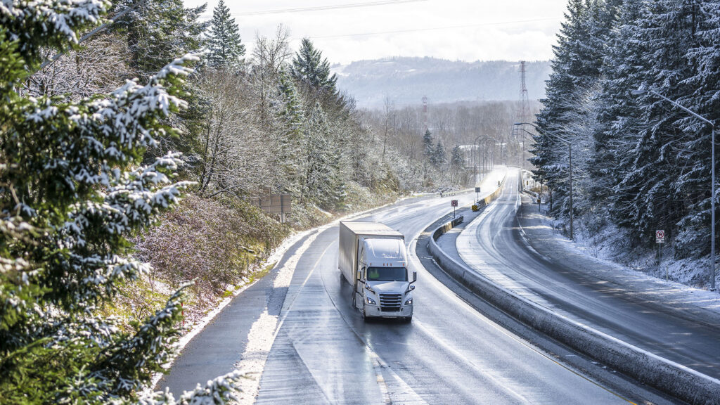 Белый современный капот, популярный профессиональный большой полуприцеп с полуприцепом сухого фургона, едущий по мокрой опасной скользкой ледяной зимней дороге со снегом на деревьях по бокам разделенного шоссе