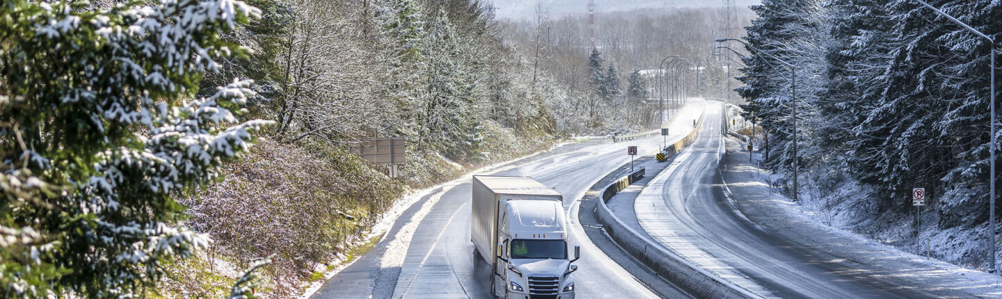 White modern bonnet popular professional big rig semi truck with dry van semi trailer going on the wet dangerous slippery icy winter road with snow on the trees on the sides of the divided highway
