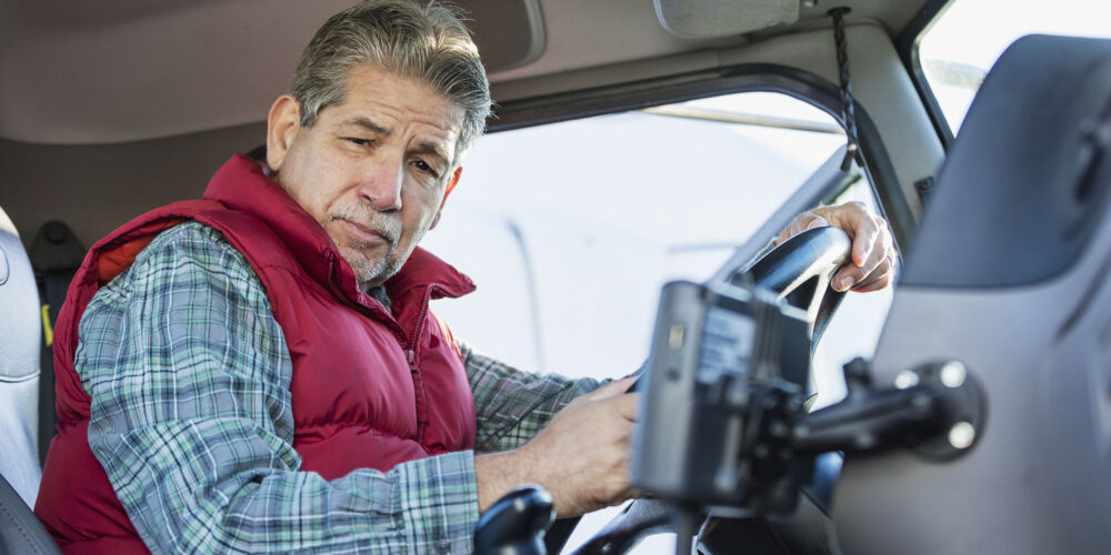 60 साल का एक वरिष्ठ हिस्पैनिक व्यक्ति सेमी-ट्रक की ड्राइवर सीट पर बैठा गंभीर भाव से कैमरे की ओर देख रहा है।