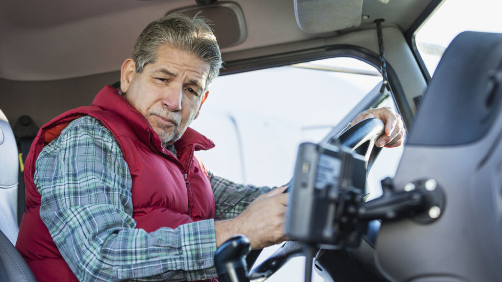 A senior Hispanic man in his 60s sitting in the driver's seat of a semi-truck, looking at the camera with a serious expression.